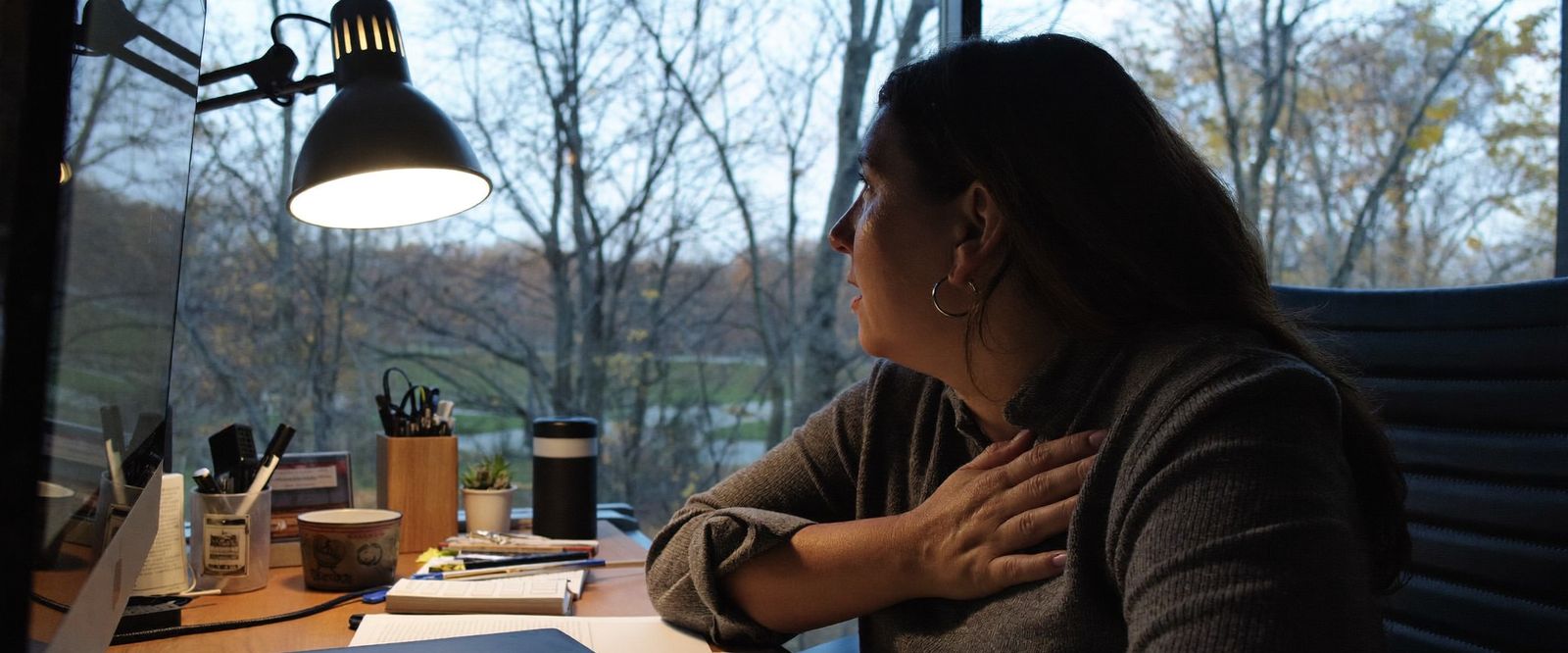 Professional woman in Michigan sitting at her home desk in the evening, hand pressed to her chest, looking exhausted yet unable to relax