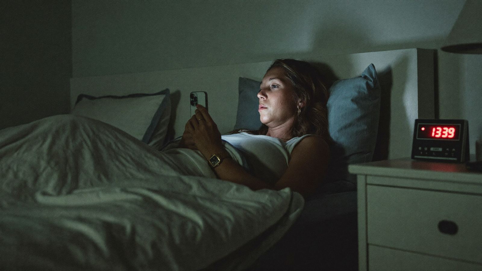 Woman awake in bed late at night in a Midwest home, phone screen lighting her face, unable to fall back asleep