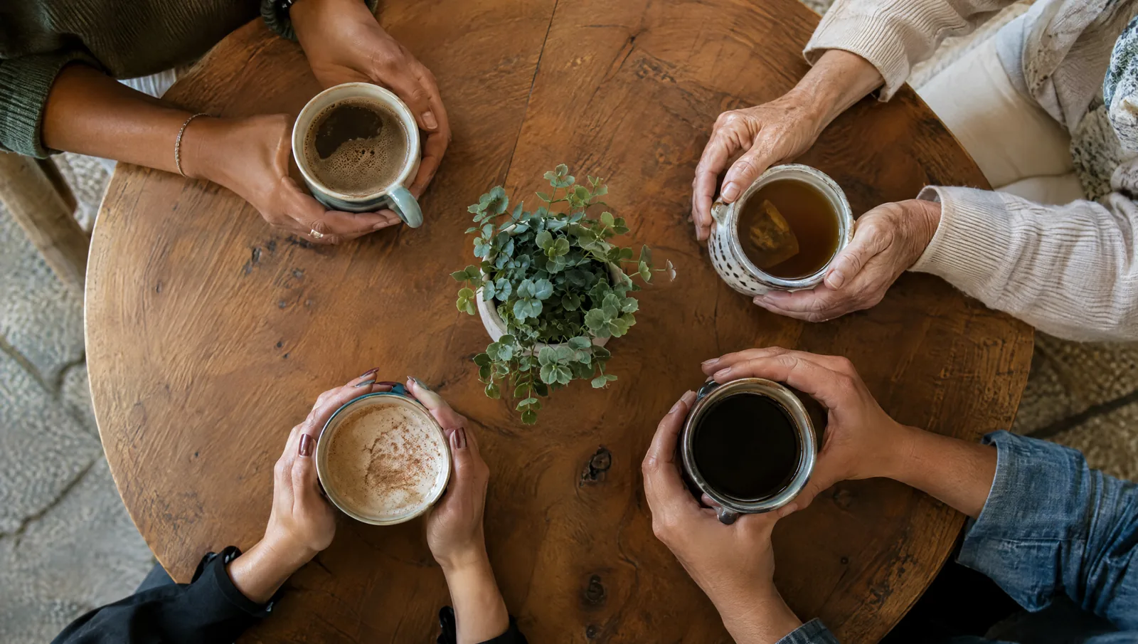 Top-down view of four women's hands of different ages and skin tones holding mugs around a round wooden table