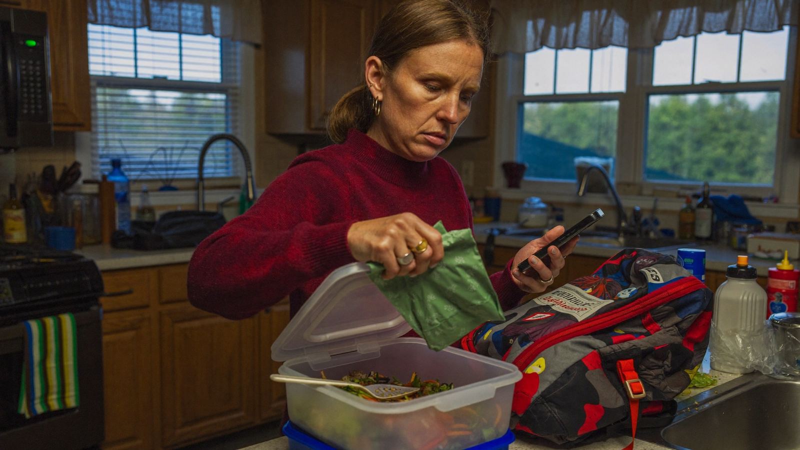 Woman in a Midwest kitchen in the early morning packing a school lunch while checking her phone, managing both childcare and work demands at once